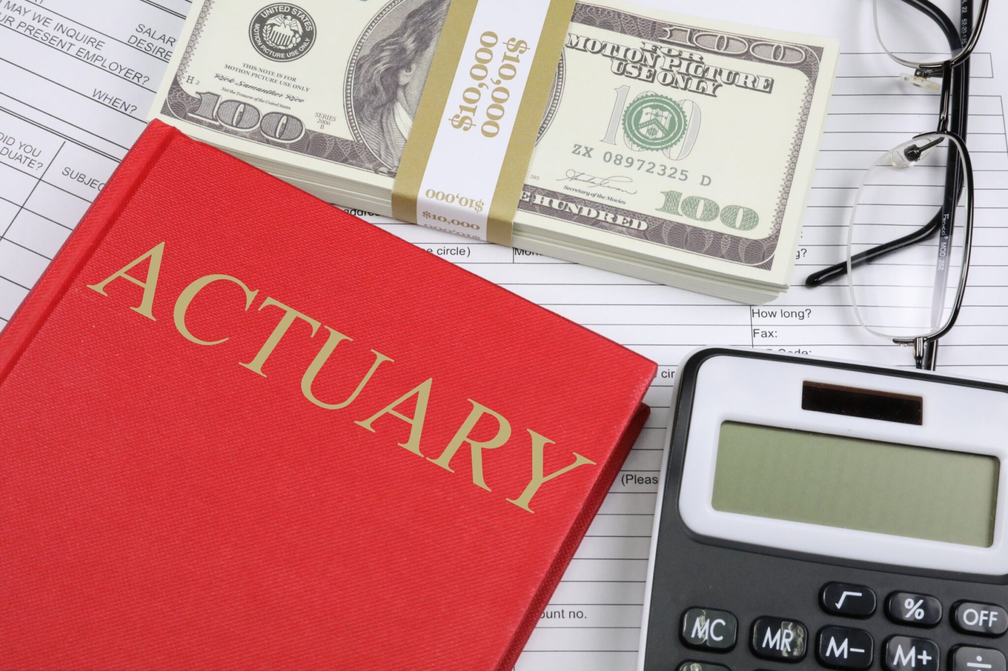 a desk of an Actuary with a calculator, some money, and a red book having actuary written on it.